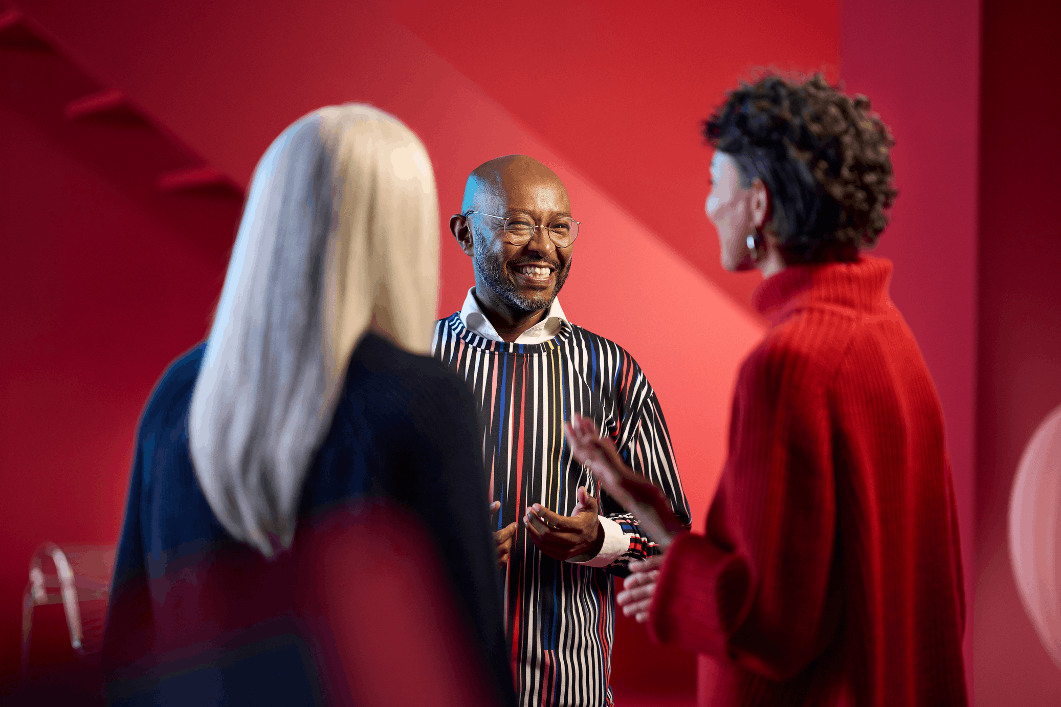 A smiling man wearing glasses and a striped sweater chatting with two women in front of a red background
